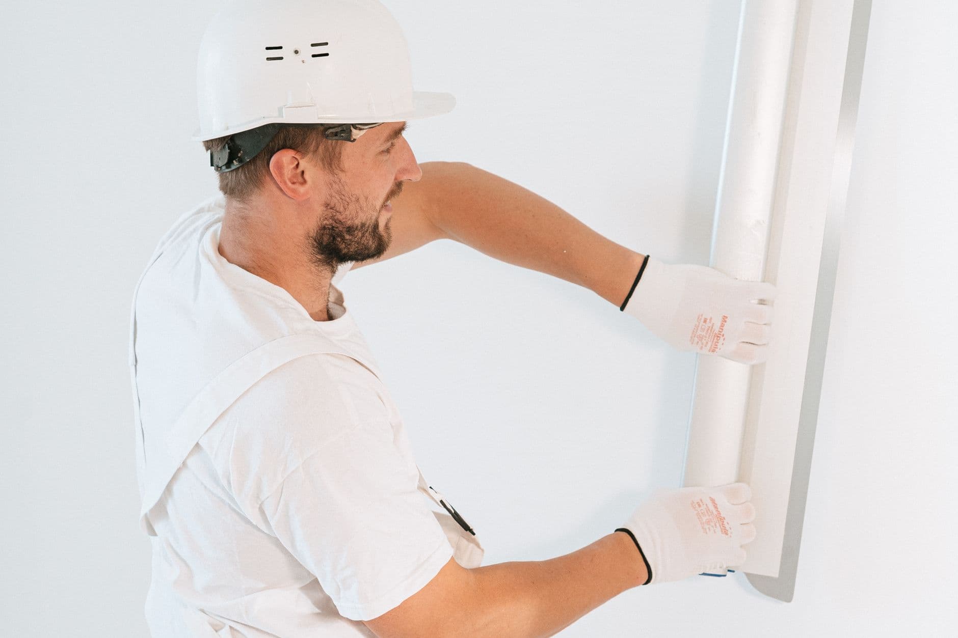 Man met witte helm, witte werkkleding en handschoenen houdt een brede spaan tegen een witte muur