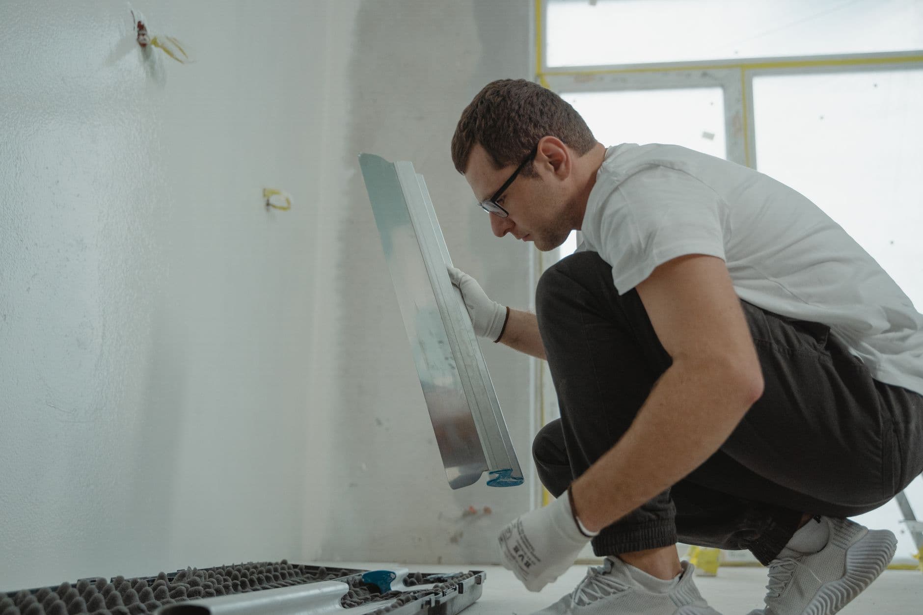 Man zit gehurkt en houdt met werkhandschoenen een lange metalen afwerkspaan bij een witte binnenmuur